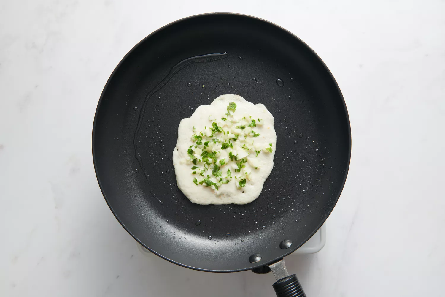 Uma frigideira antiaderente com uma pequena porção de massa no centro, coberta com cebolas picadas, pimentas e coentro